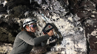 Person wearing a helmet, headlamp, and protective goggles, holding a portable science instrument up to a cave wall